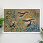 A framed vintage tapestry or wall hanging depicting three flying flamingos among detailed green, yellow, and red foliage, displayed on a white paneled wall with small green plants at the sides.