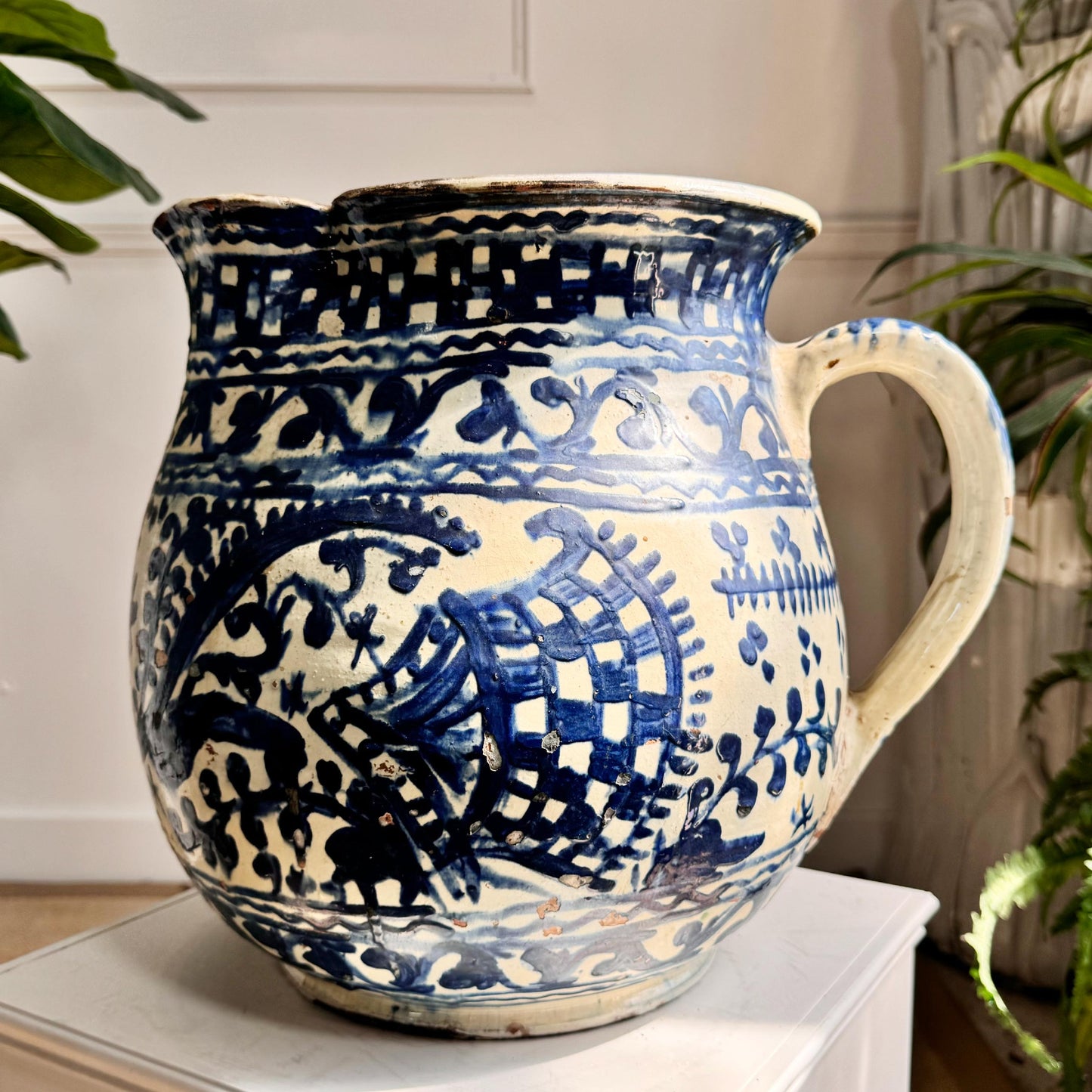 Antique Troita Popa Slipware Jug Romanian 1920's with a blue and cream floral and scroll pattern, displayed on a white pedestal in an indoor setting with plants in the background.