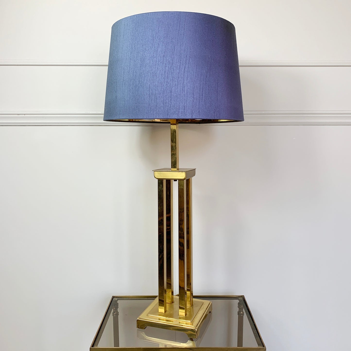A designer table lamp with a textured blue-gray fabric drum shade and a polished gold-finished metal base, displayed on a mirrored side table against a white paneled wall.