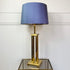 A designer table lamp with a textured blue-gray fabric drum shade and a polished gold-finished metal base, displayed on a mirrored side table against a white paneled wall.
