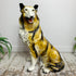 A glossy ceramic figurine of a collie dog sitting on a rustic wooden floor against a plain white wall, with green fern plants visible on both sides.