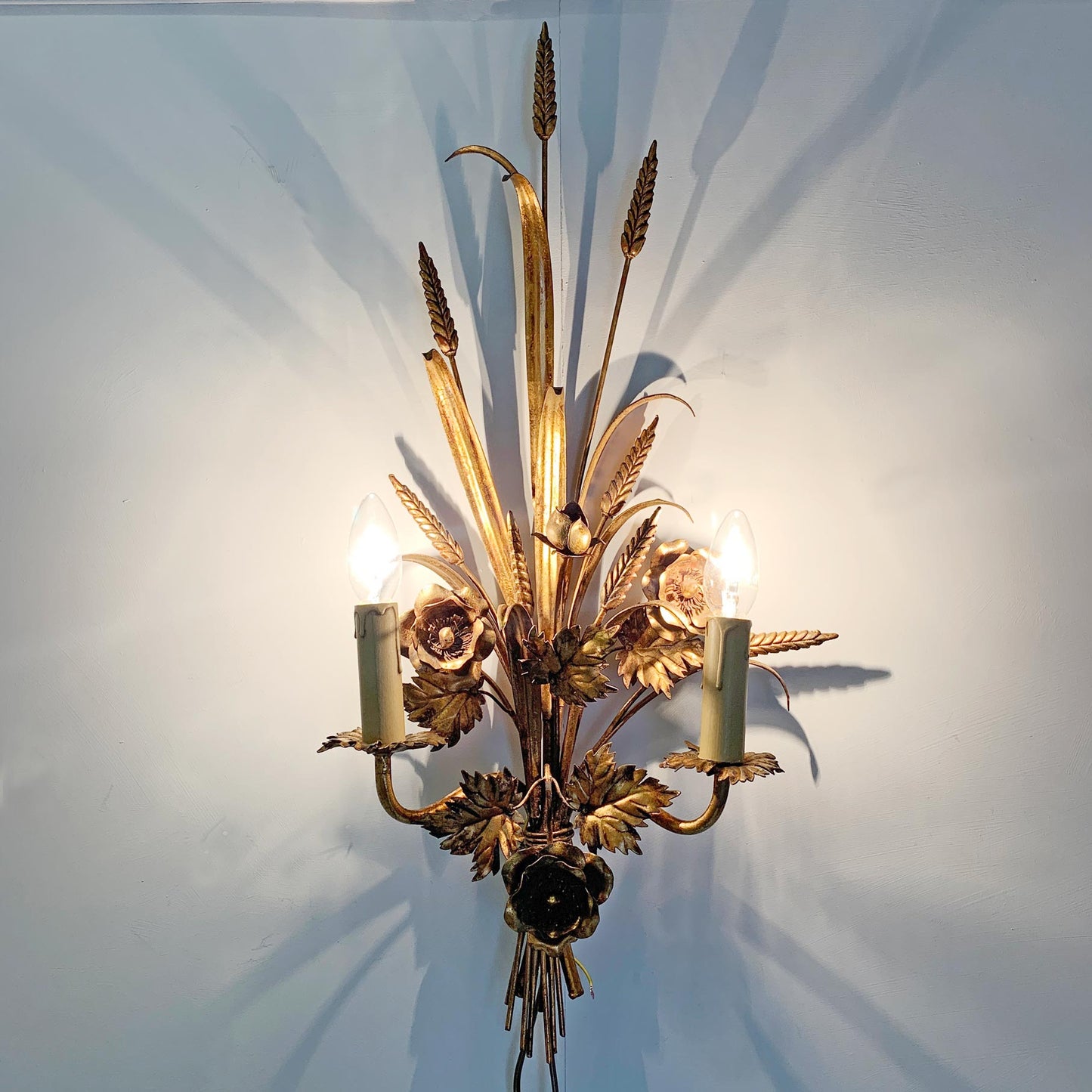 A decorative wall sconce with a warm gold finish, featuring wheat stalks, leaves, and candle-style bulbs, mounted on a light-colored wall.