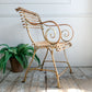 A wrought iron Arras garden chair with scrollwork armrests and a slatted back and seat, shown on a rustic wood floor next to a potted fern against a white paneled wall.