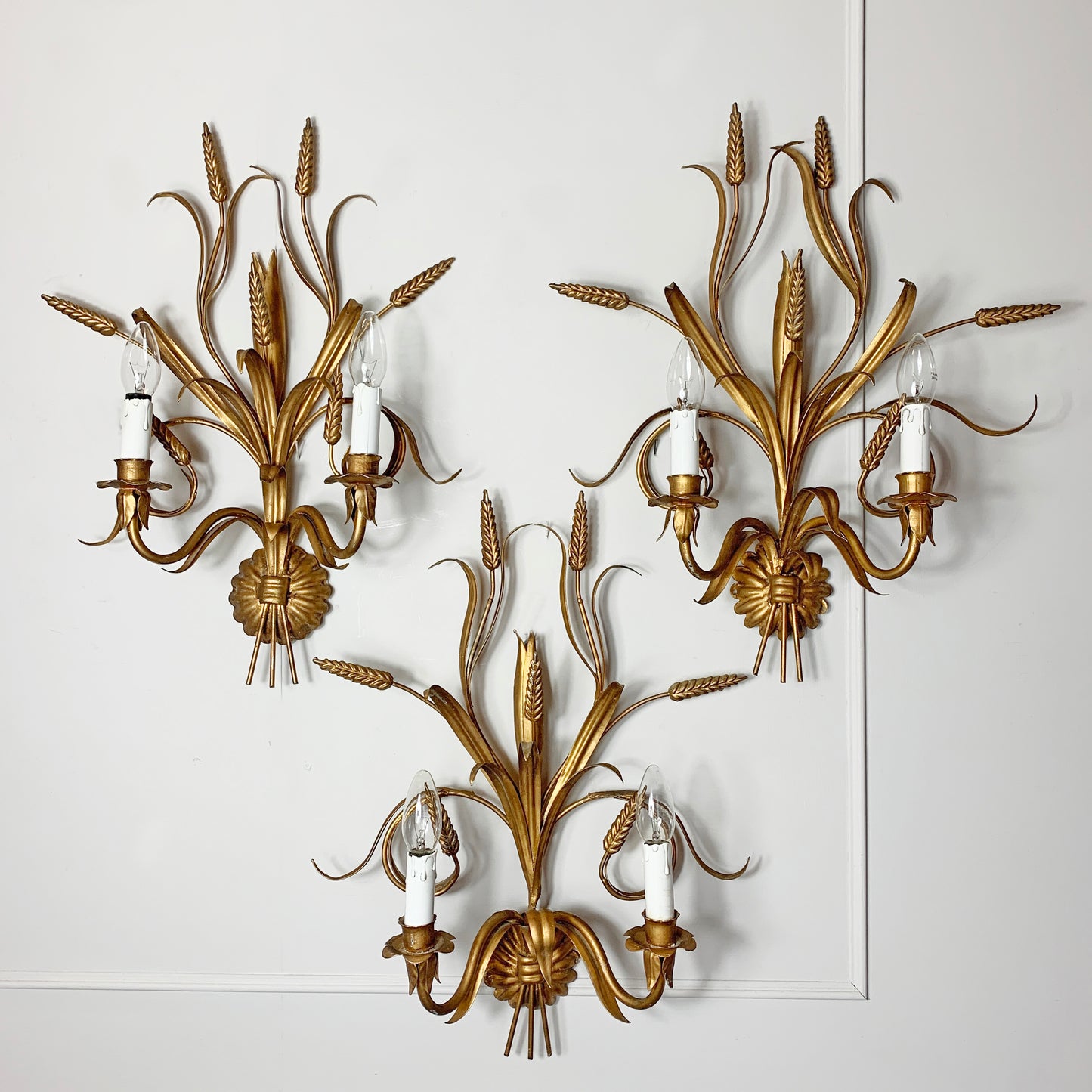 Three ornate Hans Kogl gold wall sconces with a wheat or reed-inspired leaf design, each holding two candle-style bulbs, mounted on a white paneled wall.