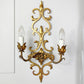 An ornate gold metal two-light wall sconce with intricate scrollwork and candle-style bulbs, mounted on a white paneled wall.