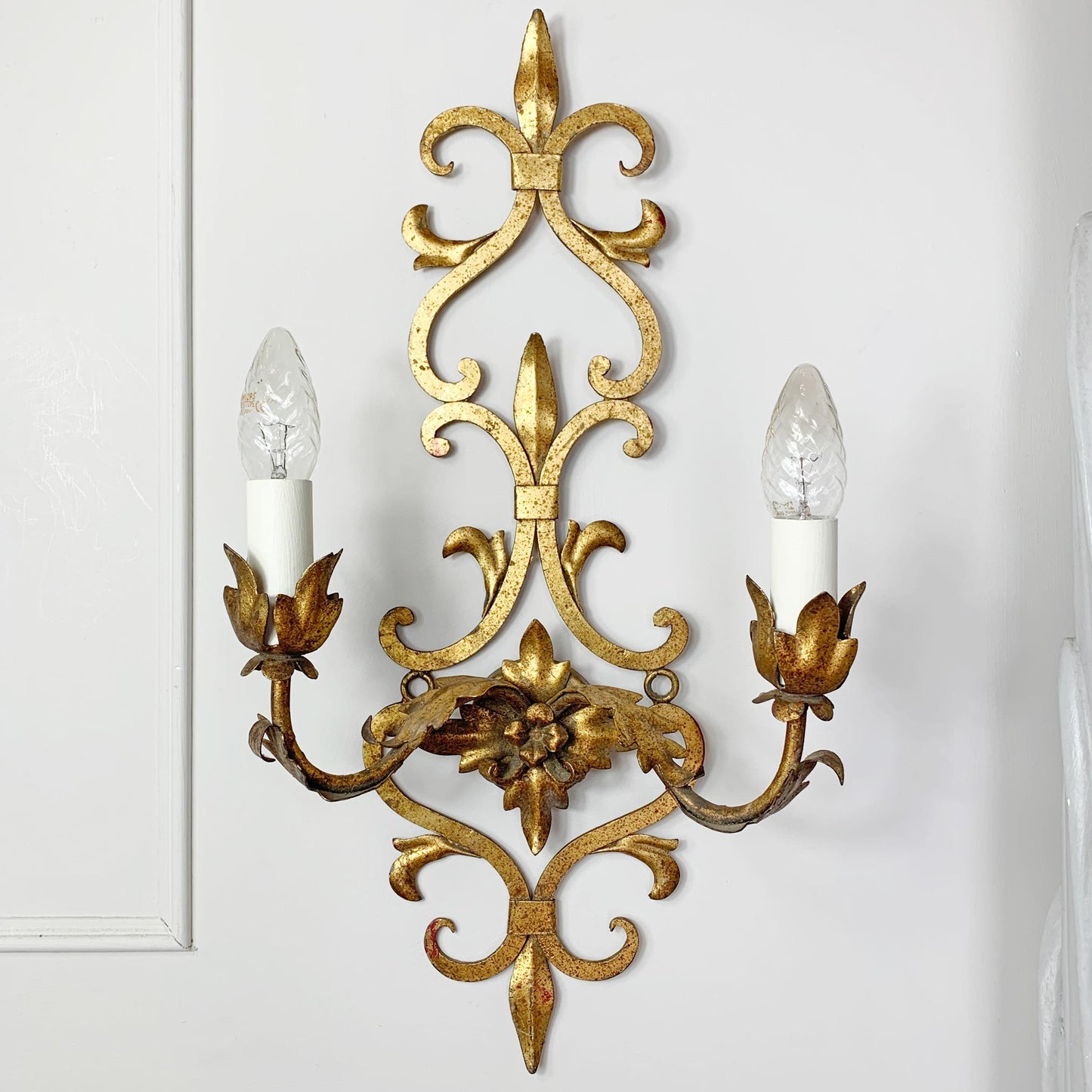 An ornate gold metal two-light wall sconce with intricate scrollwork and candle-style bulbs, mounted on a white paneled wall.