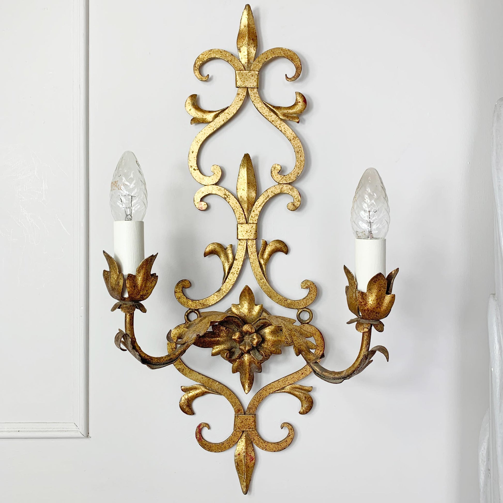 An ornate gold metal two-light wall sconce with intricate scrollwork and candle-style bulbs, mounted on a white paneled wall.