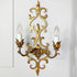 An ornate gold metal two-light wall sconce with intricate scrollwork and candle-style bulbs, mounted on a white paneled wall.