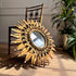 A decorative sunburst wall mirror with a gold-toned frame and leaf-like rays, leaning against a wooden chair in an indoor room with a plant in the background.