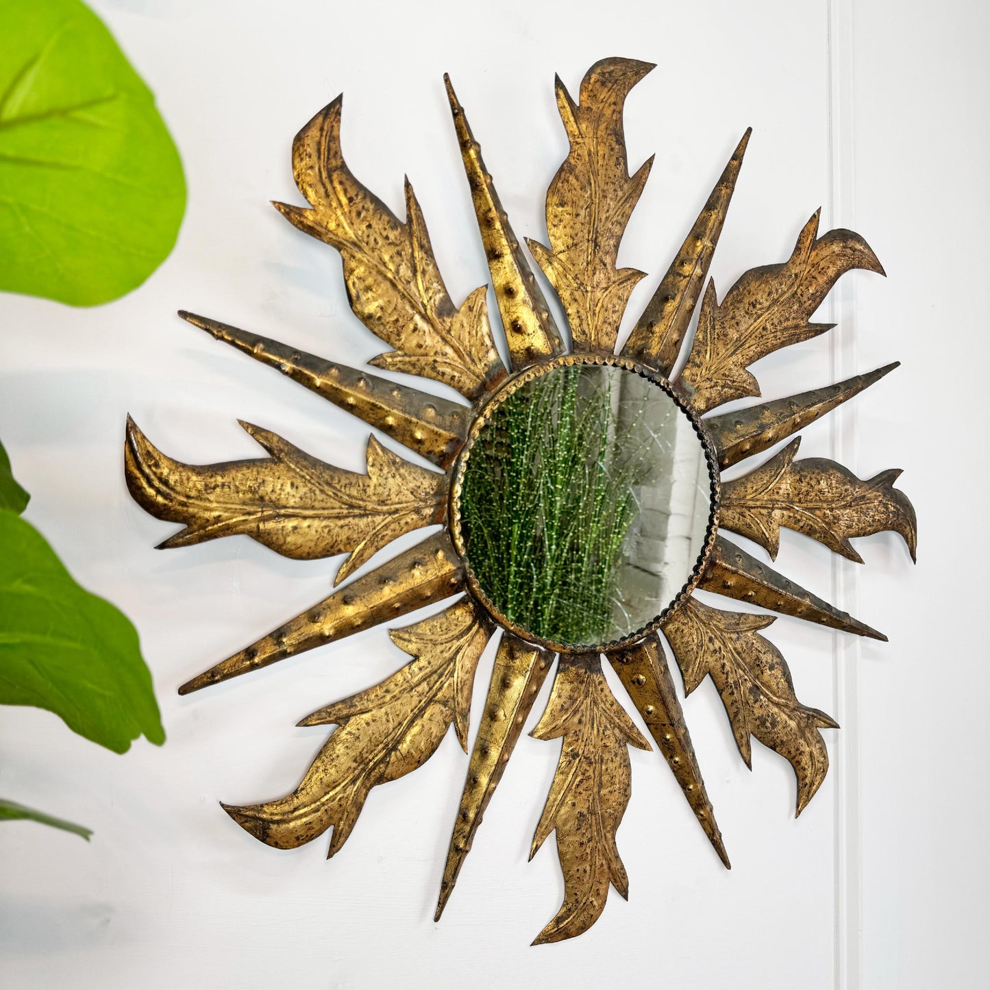 A decorative sunburst wall mirror with a distressed gold metal frame and leaf-like rays, mounted on a white wall with green leaves visible at the left edge.