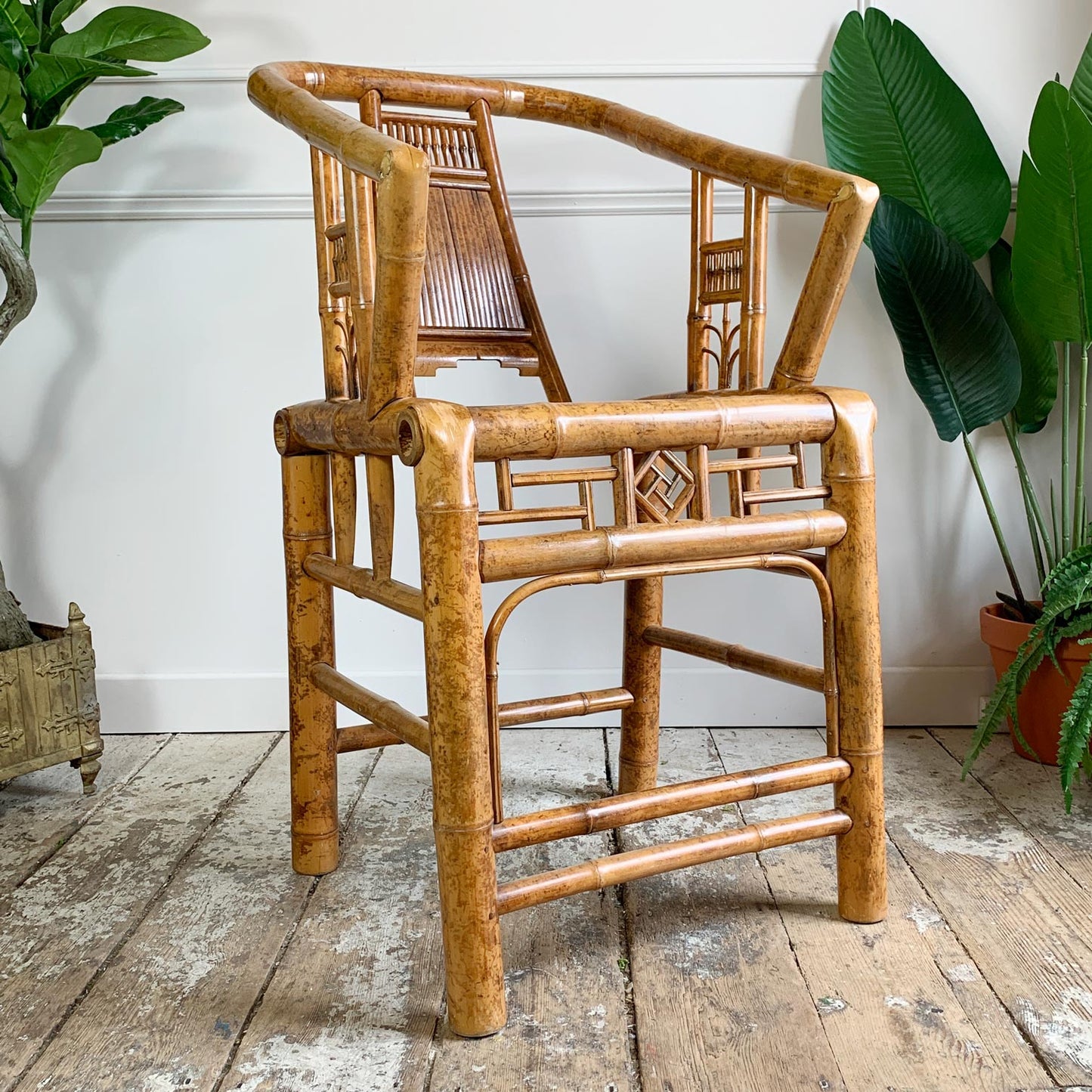 A natural bamboo armchair with a woven cane backrest, set on a rustic wooden floor against a white wall with potted plants in the background.