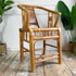 A natural bamboo armchair with a woven cane backrest, set on a rustic wooden floor against a white wall with potted plants in the background.