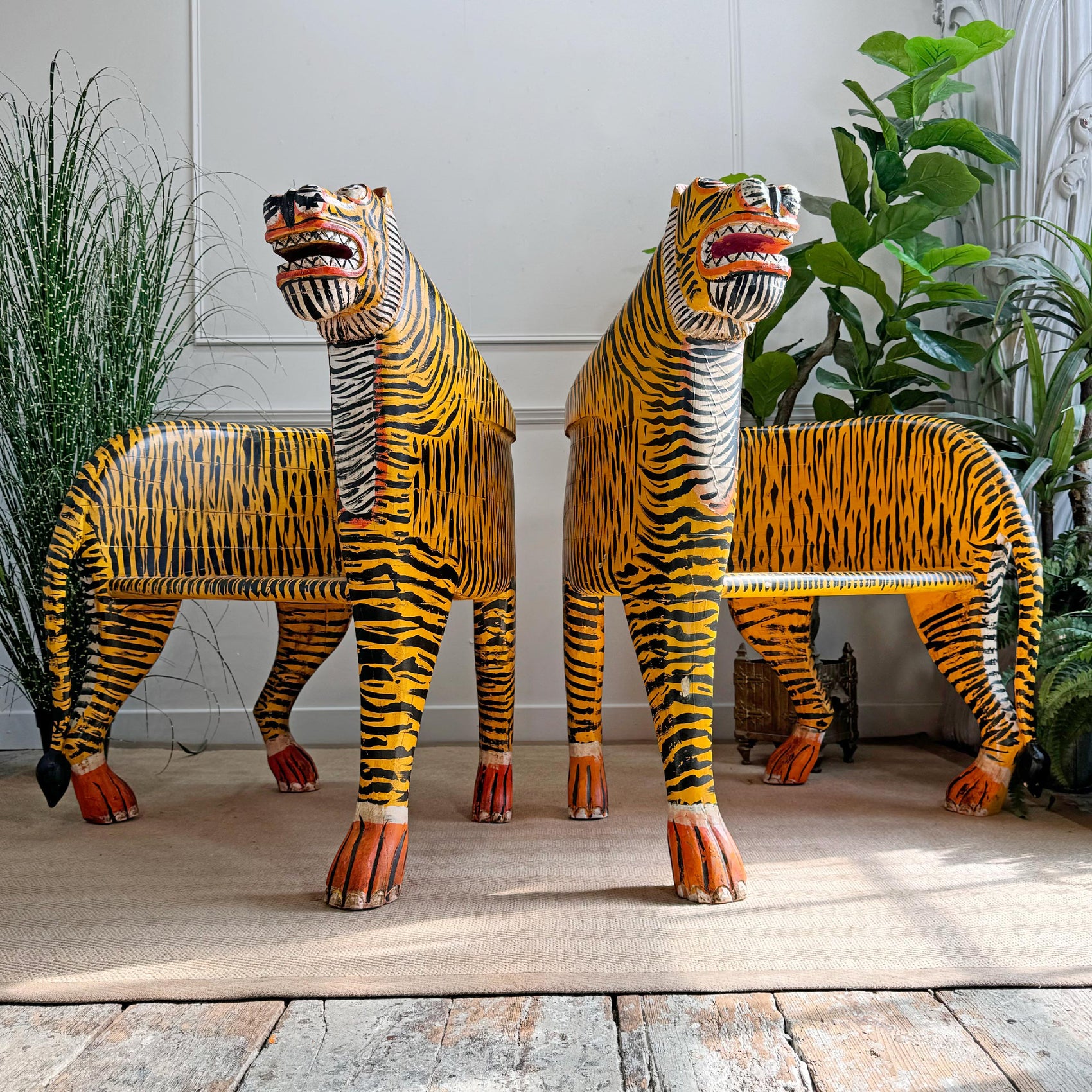 Two hand-painted wooden tiger sculptures or chairs with yellow and black stripes, displayed in a styled indoor setting with plants and a rustic wooden floor.