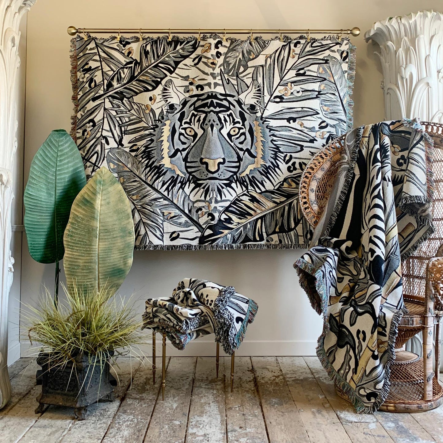 A large woven tapestry featuring a stylized tiger face surrounded by tropical leaves, displayed in a styled room setting with matching textiles, plants, and rattan furniture.