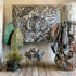 A large woven tapestry featuring a stylized tiger face surrounded by tropical leaves, displayed in a styled room setting with matching textiles, plants, and rattan furniture.