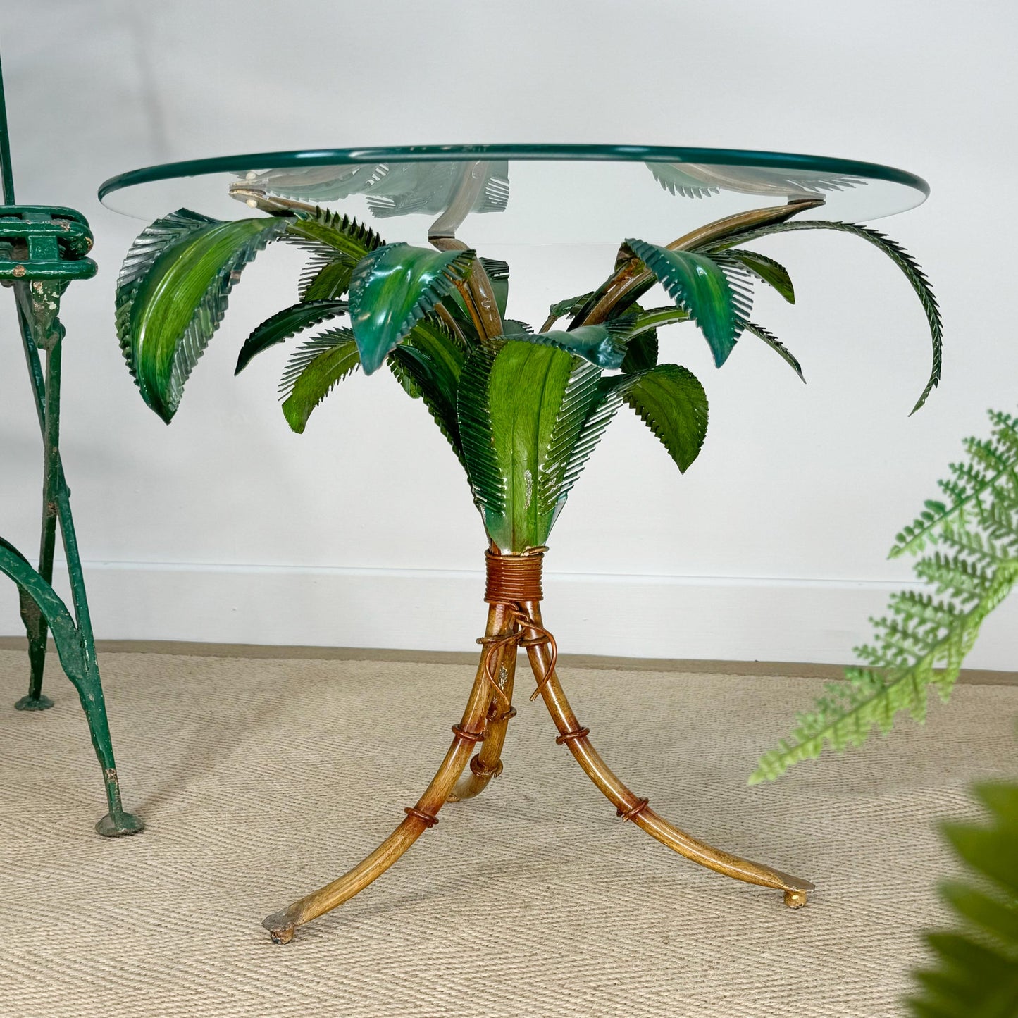 A tropical-themed side table featuring a clear glass top, a green faux palm tree metal frame, and a gold-finished tripod base with faux rattan details, set on a beige carpeted floor.