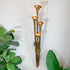 A vintage brass wall sconce with four tiered trumpet-shaped candle holders, lit with warm glowing candles, mounted on a white paneled wall with green houseplants in the foreground.