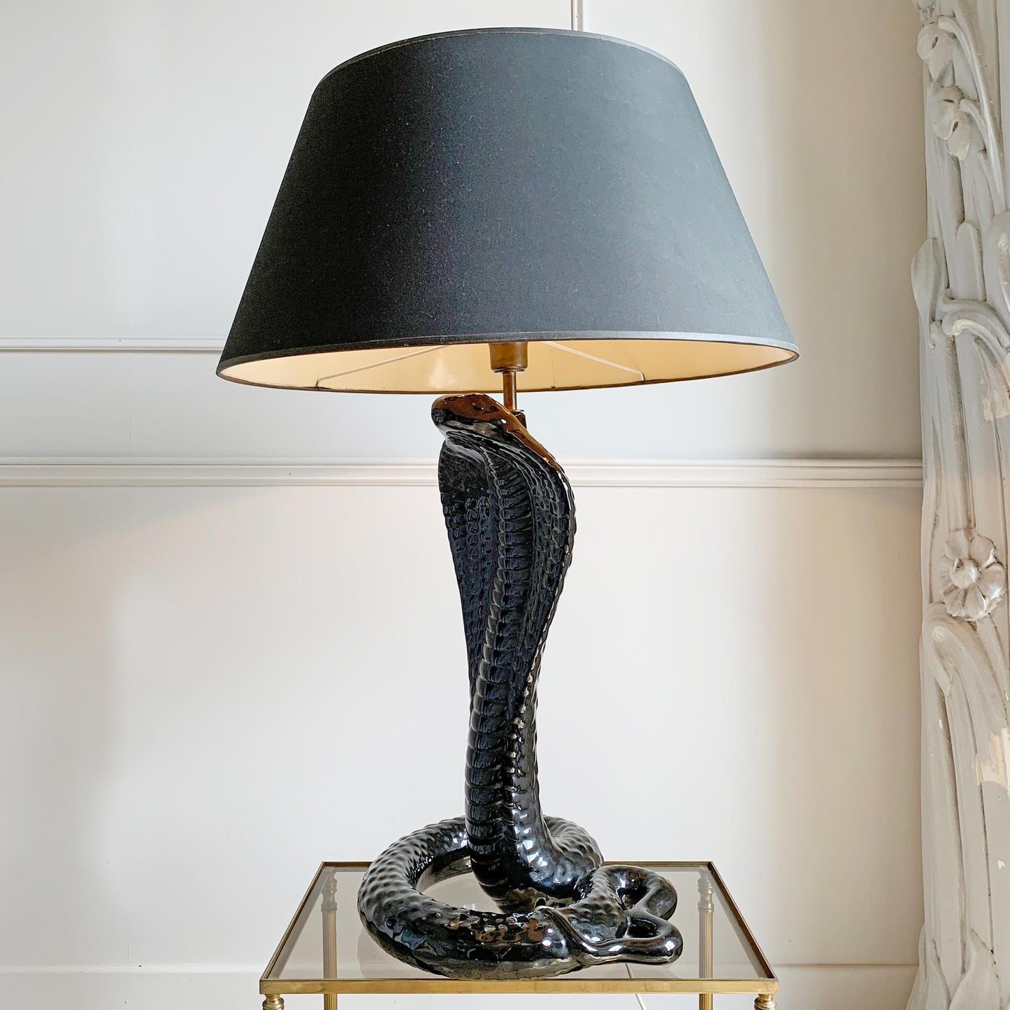 A tomasso barbi vintage table lamp featuring a sculpted black ceramic cobra snake base and a large black drum shade with a warm gold interior, displayed on a glass-topped side table against a cream wall.