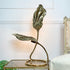 An Tommaso Barbi Gold Double Leaf metal table lamp shaped like stylized leaves on a curved stem, standing on a round base. The lamp has a polished metallic finish in a warm bronze or gold tone and is photographed indoors on a glass-topped side table against a white paneled wall.