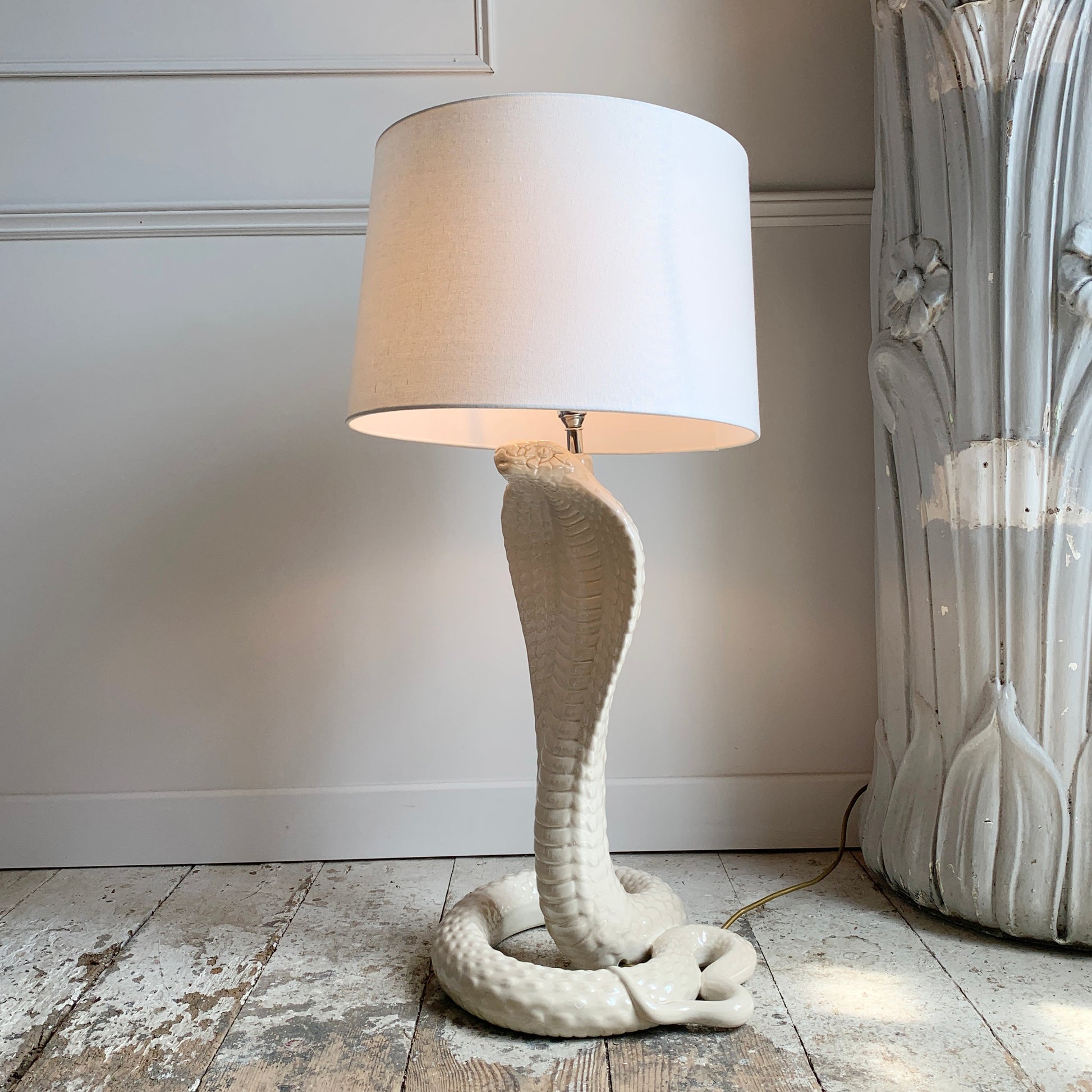 A Tommaso Barbi table lamp shaped like a coiled cobra snake in an off-white ceramic, with a matching off-white drum shade, set on a rustic wooden floor against a white paneled wall.