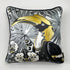 A square decorative silk pillow featuring a stylized toucan illustration in black, white, gray, and gold, with a black piped edge, shown against a light gray background.
