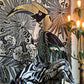 A large woven tapestry featuring a stylized toucan and tropical floral motifs in black, gray, white, and gold, draped over a chair in an interior setting with a lit floor lamp visible on the right.
