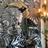 A large woven tapestry featuring a stylized toucan and tropical floral motifs in black, gray, white, and gold, draped over a chair in an interior setting with a lit floor lamp visible on the right.