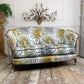 An antique loveseat sofa with a white fabric upholstered in a bold yellow and gray palm leaf pattern, featuring a natural wood frame and legs, set against a white paneled wall with a brass wall sconce.