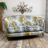 An antique loveseat sofa with a white fabric upholstered in a bold yellow and gray palm leaf pattern, featuring a natural wood frame and legs, set against a white paneled wall with a brass wall sconce.