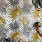 A close-up of a wallpaper pattern featuring stylized palm fronds in metallic gold and matte gray on a white background, with decorative props like a bamboo lamp base and textured coral at the bottom.