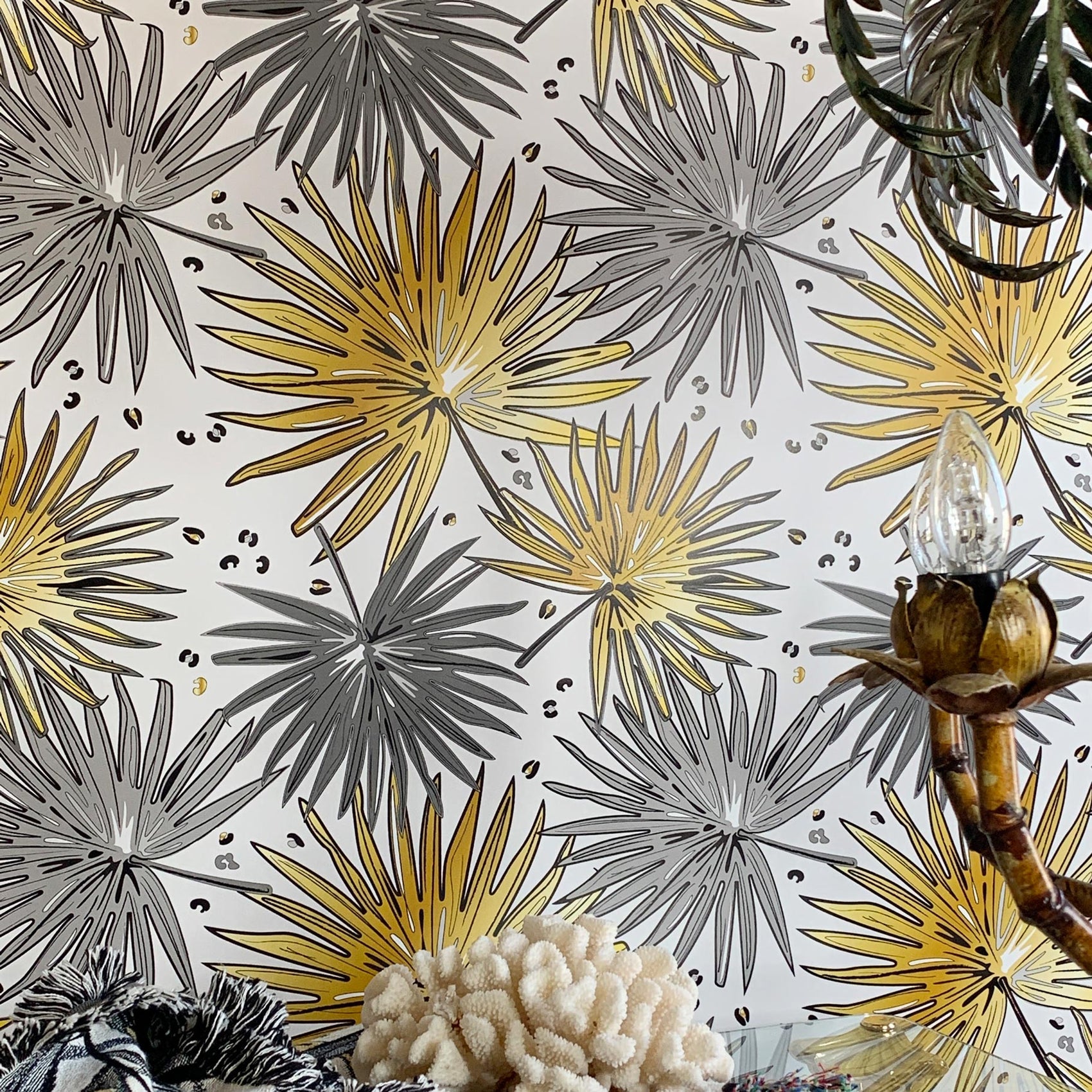 A close-up of a wallpaper pattern featuring stylized palm fronds in metallic gold and matte gray on a white background, with decorative props like a bamboo lamp base and textured coral at the bottom.