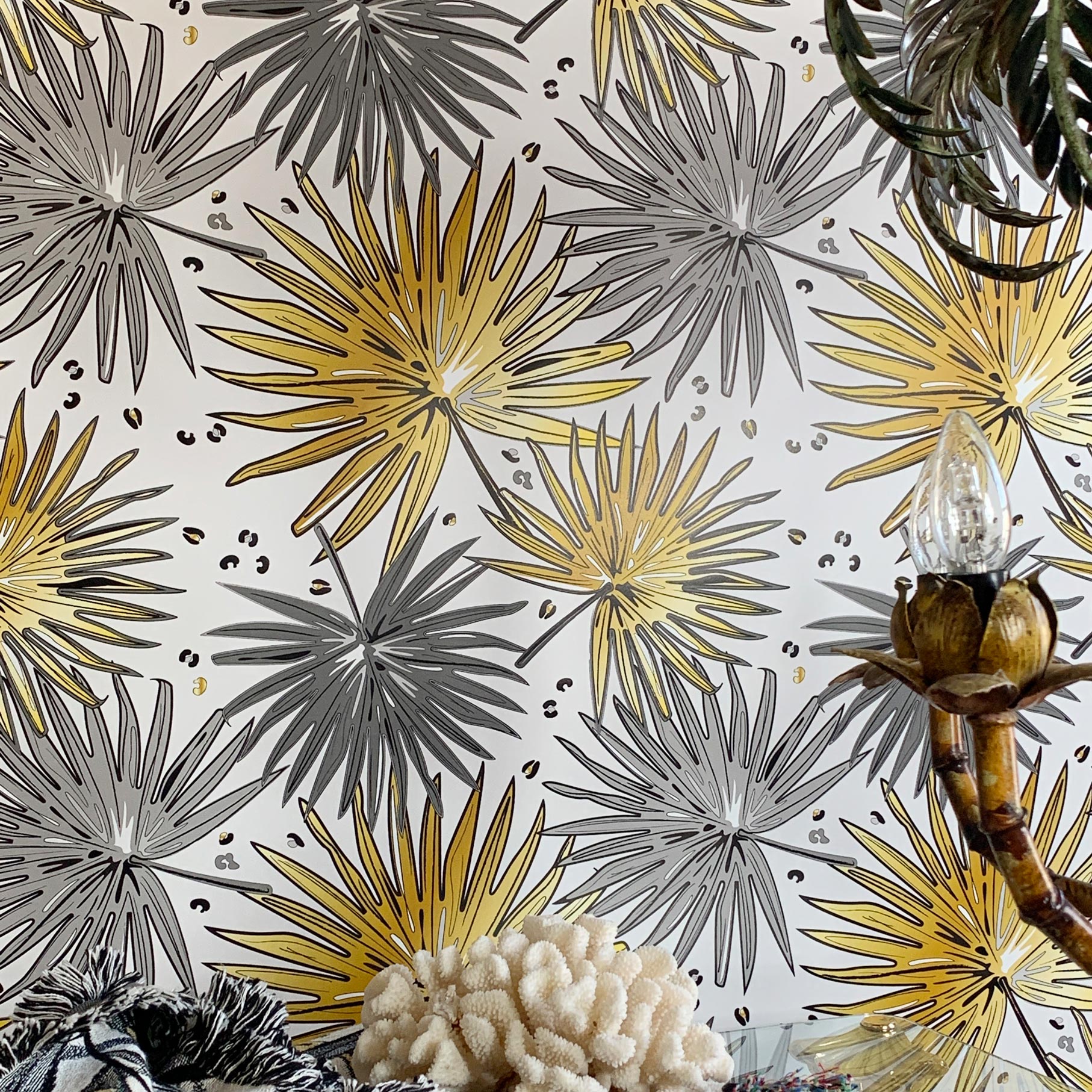 A close-up of a wallpaper pattern featuring stylized palm fronds in metallic gold and matte gray on a white background, with decorative props like a bamboo lamp base and textured coral at the bottom.