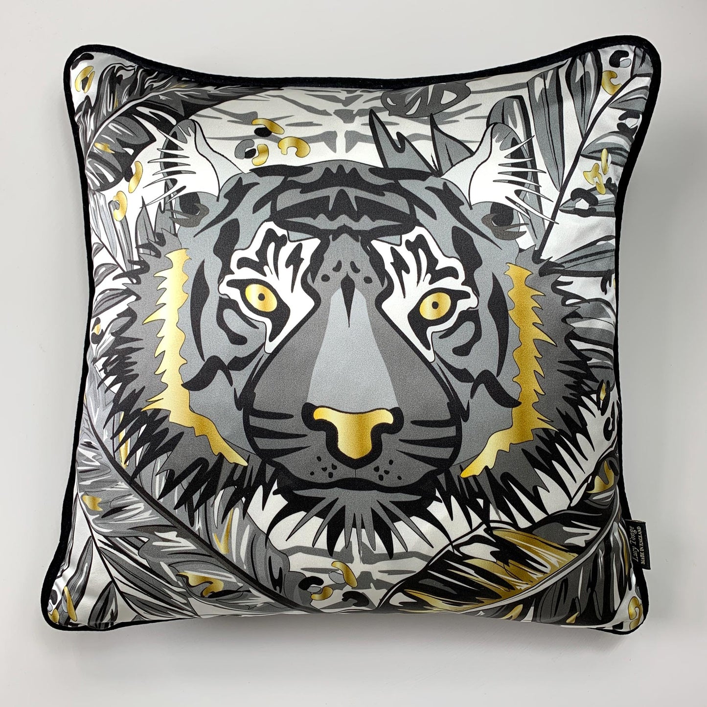 A square decorative silk pillow featuring a stylized tiger face illustration in black, gray, white, and gold, with a black piped edge and a small black brand tag on the right side.