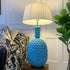A large table lamp with a bright blue pineapple-shaped ceramic base and a white pleated drum shade, shown lit in a styled interior setting next to a sofa and houseplant.