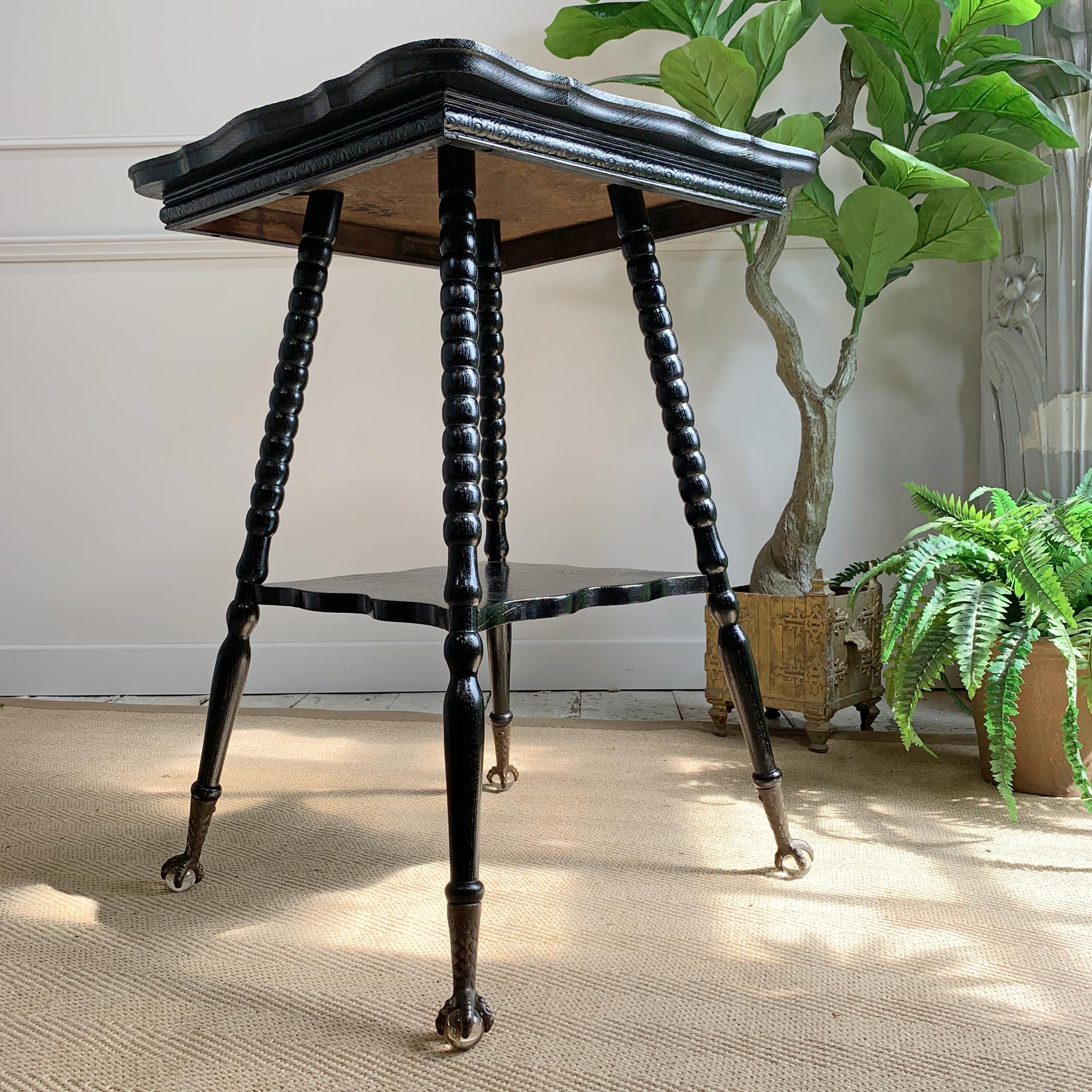A black ebonised wooden corner table with turned legs and brass-toned claw casters, photographed in a styled interior setting with potted plants and a light wall background.