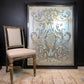 A large rectangular framed wall pub glass mirror with an ornate silver damask pattern on a distressed silver and gold background, leaning against a dark wall next to a vintage wooden chair.