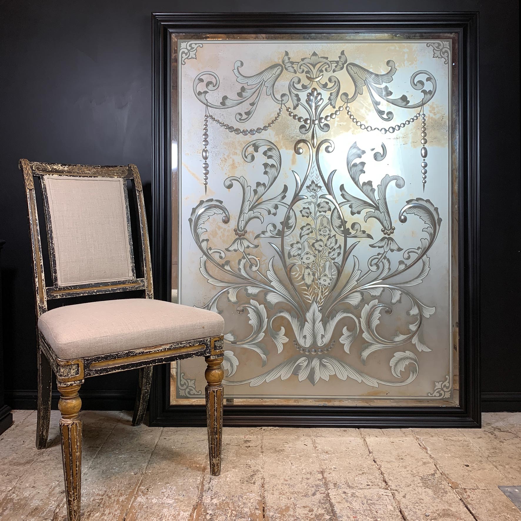 A large rectangular framed wall pub glass mirror with an ornate silver damask pattern on a distressed silver and gold background, leaning against a dark wall next to a vintage wooden chair.