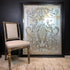A large rectangular framed wall pub glass mirror with an ornate silver damask pattern on a distressed silver and gold background, leaning against a dark wall next to a vintage wooden chair.