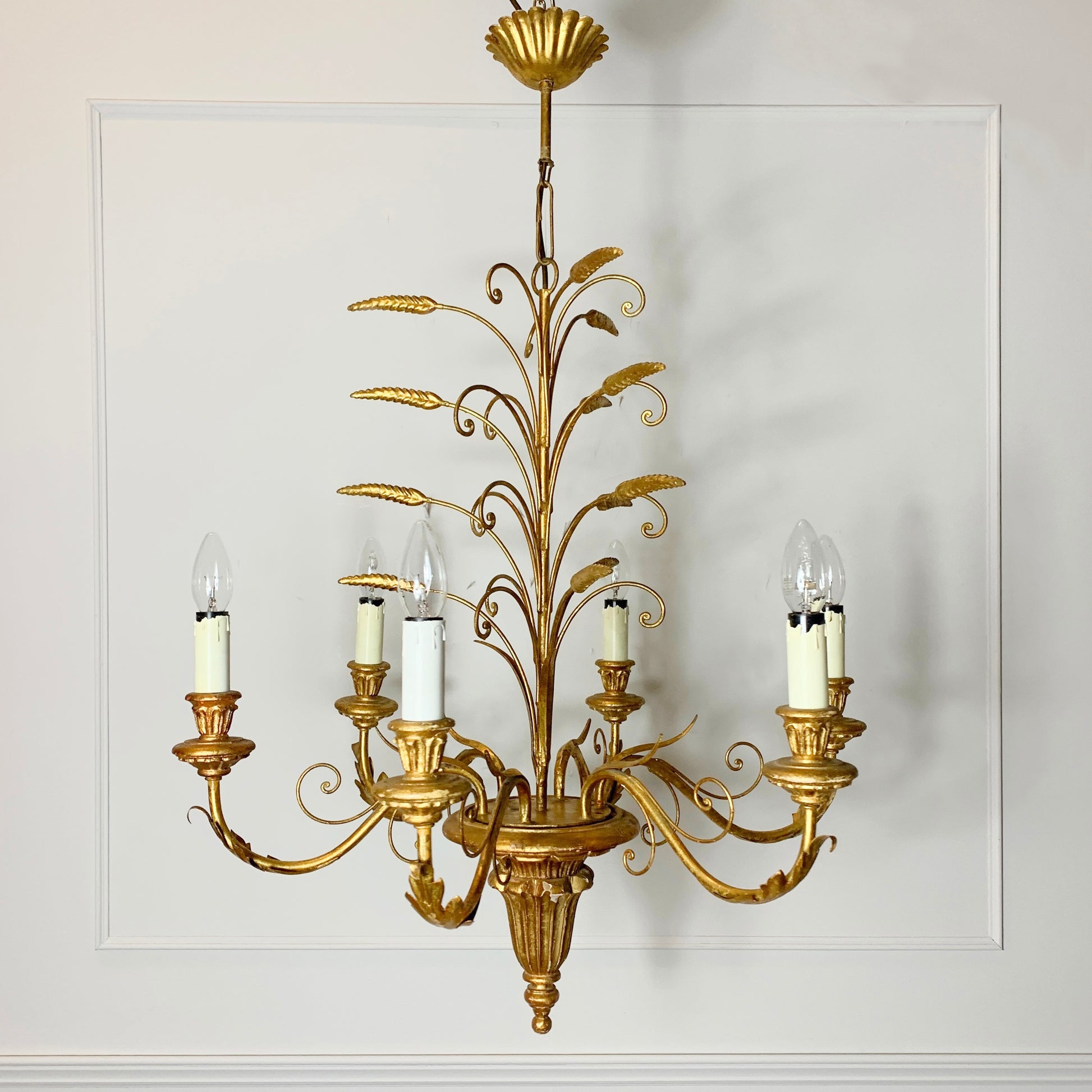 A decorative six-light wall or ceiling chandelier with an ornate gold metal frame styled like a wheatsheaf, featuring candle-style bulbs on curved arms against a white paneled wall.