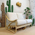 A two-seater rattan sofa with thick off-white cushions, styled in a room with decorative green cacti, a wicker planter, and a gold leaf wall piece.