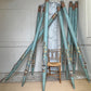 A set of long decorative teal-blue zinc metal church organ pipes with ornate painted floral and geometric motifs in gold, brown, and cream, arranged in a fan shape around a small woven-back chair in an indoor room.