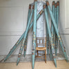 A set of long decorative teal-blue zinc metal church organ pipes with ornate painted floral and geometric motifs in gold, brown, and cream, arranged in a fan shape around a small woven-back chair in an indoor room.