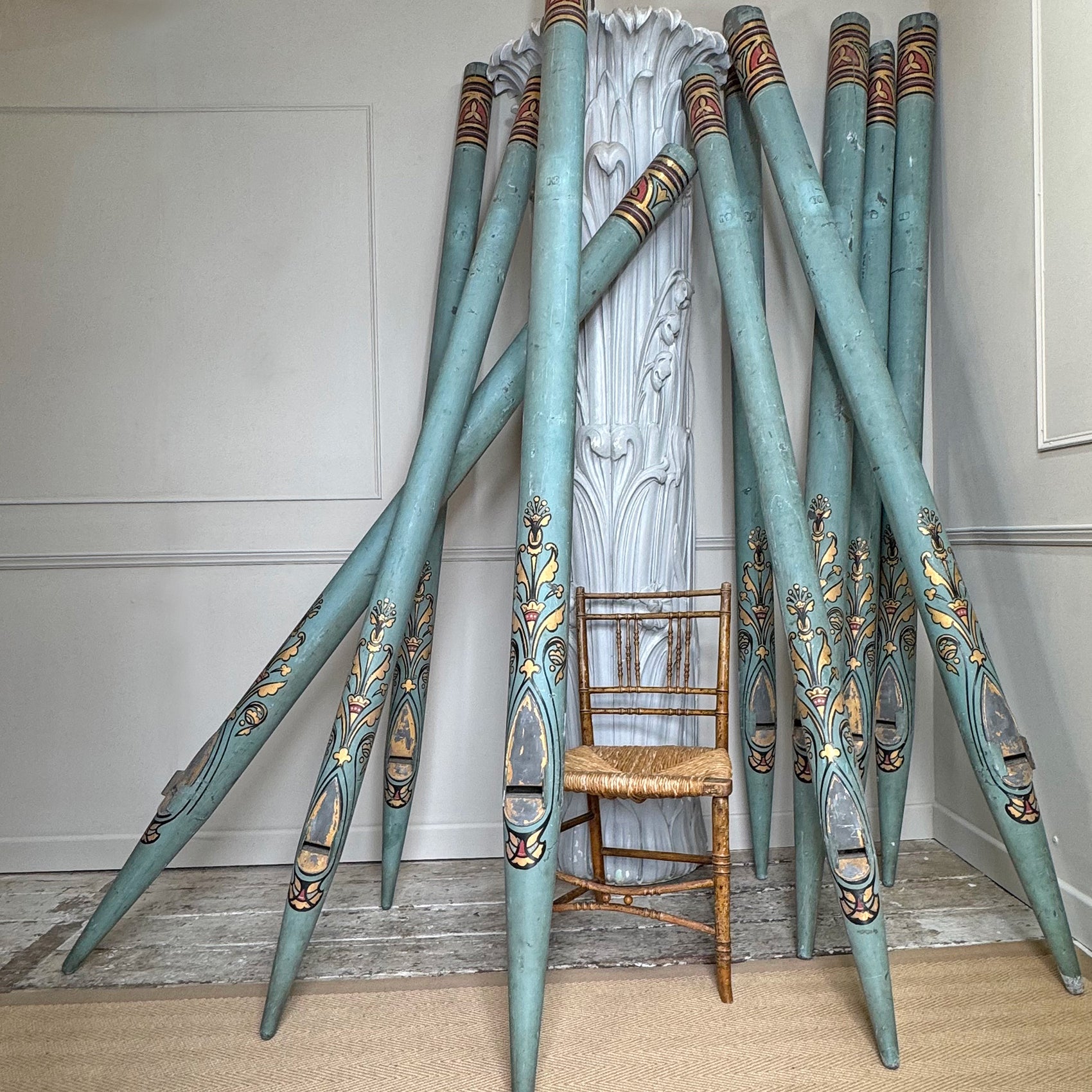 A set of long decorative teal-blue zinc metal church organ pipes with ornate painted floral and geometric motifs in gold, brown, and cream, arranged in a fan shape around a small woven-back chair in an indoor room.
