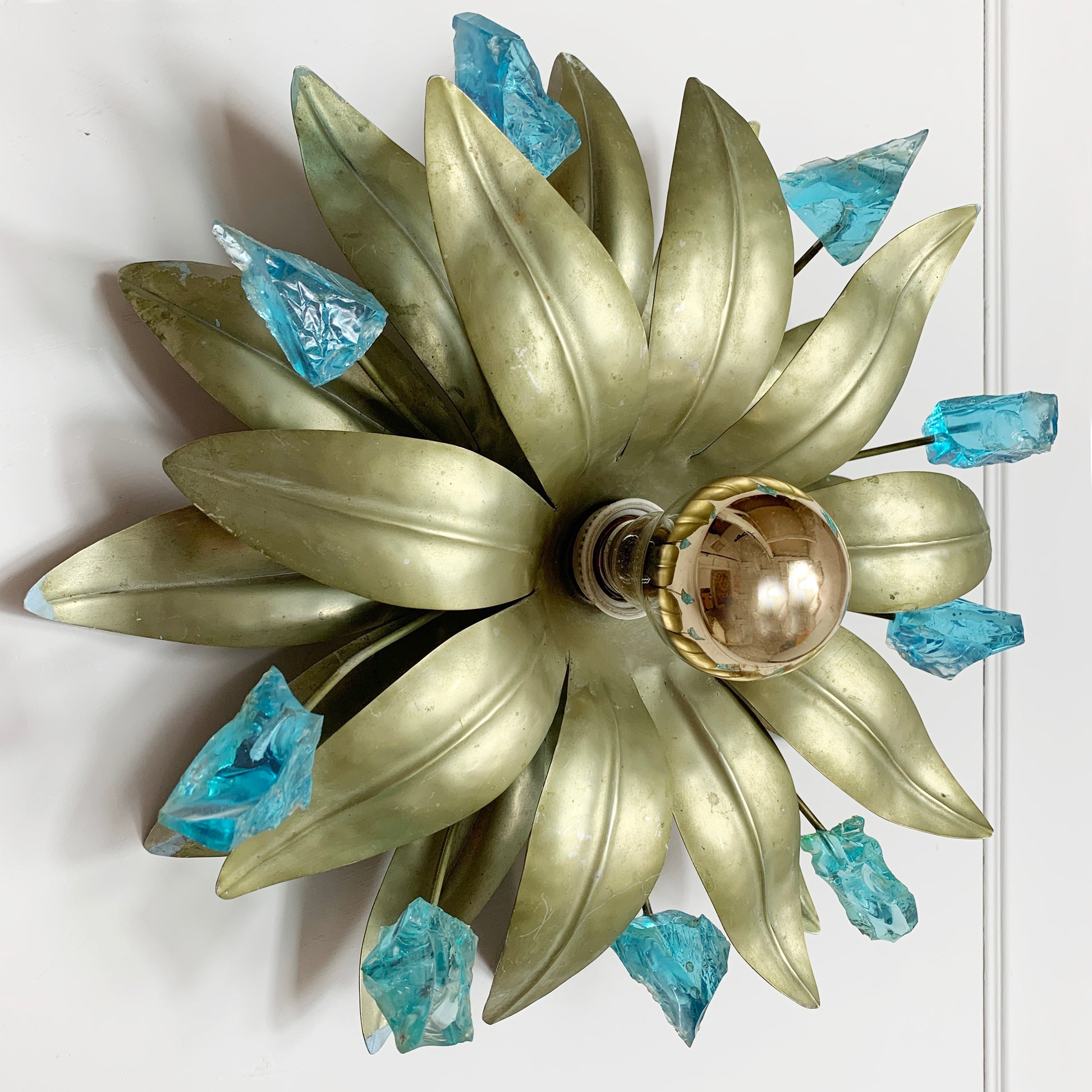 A decorative wall-mounted light fixture shaped like a sunflower, featuring metallic gold-toned petals, a clear glass globe bulb at the center, and translucent blue crystal-like accents around the outer petals, shown against a plain white wall.