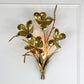 A decorative gold metallic wall sconce shaped like a floral bouquet with metallic leaves and stems, mounted on a white wall.