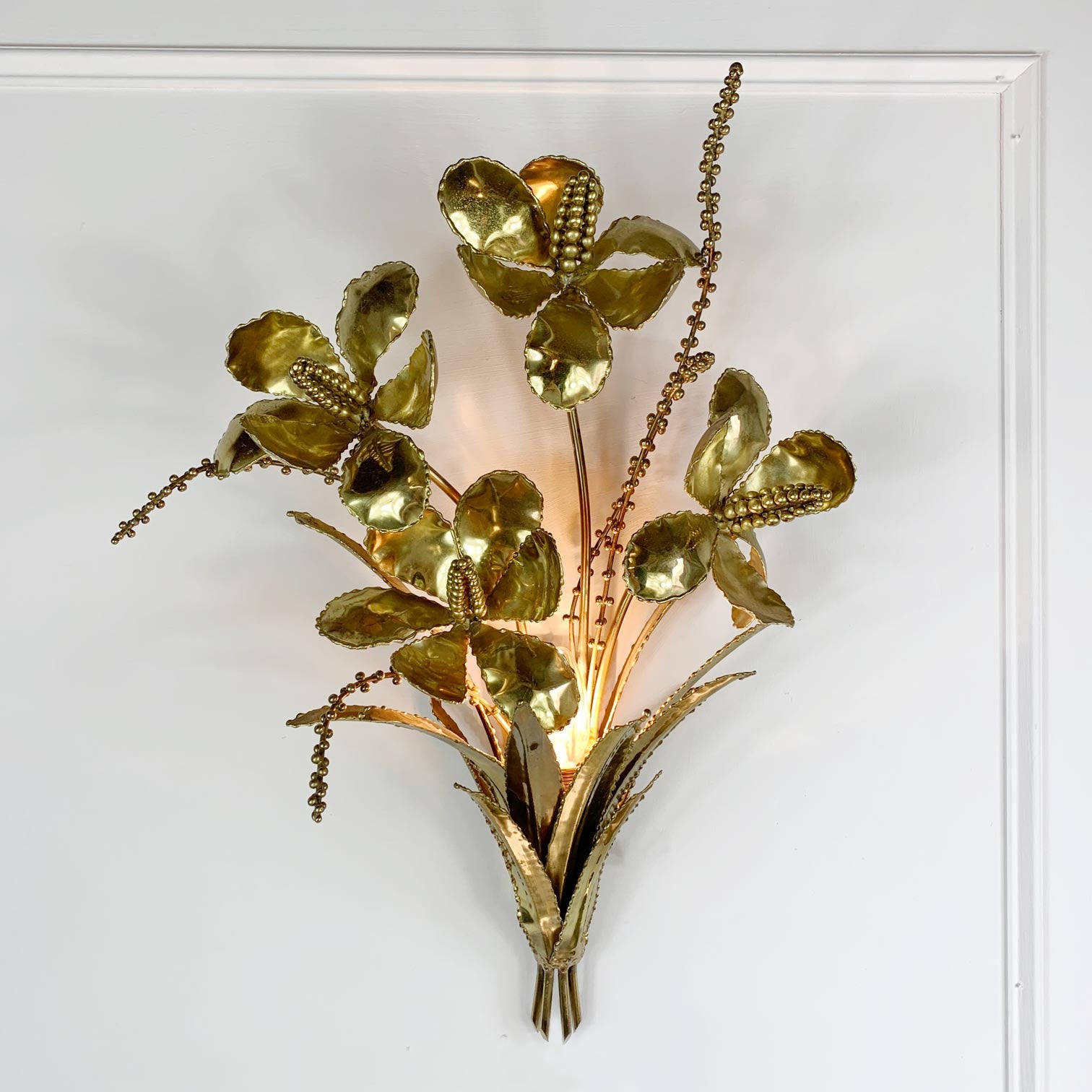 A decorative gold metallic wall sconce shaped like a floral bouquet with metallic leaves and stems, mounted on a white wall.