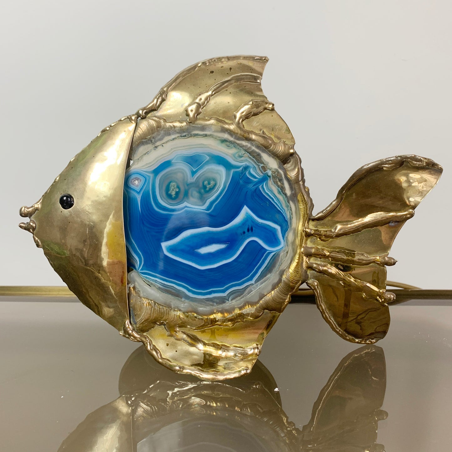 A Jacques Duval-Brasseur Illuminated Blue Agate Gold Fish shaped lamp with a textured metallic gold body and a central blue agate inlay, photographed against a plain light background with a reflective surface below.