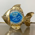 A Jacques Duval-Brasseur Illuminated Blue Agate Gold Fish shaped lamp with a textured metallic gold body and a central blue agate inlay, photographed against a plain light background with a reflective surface below.
