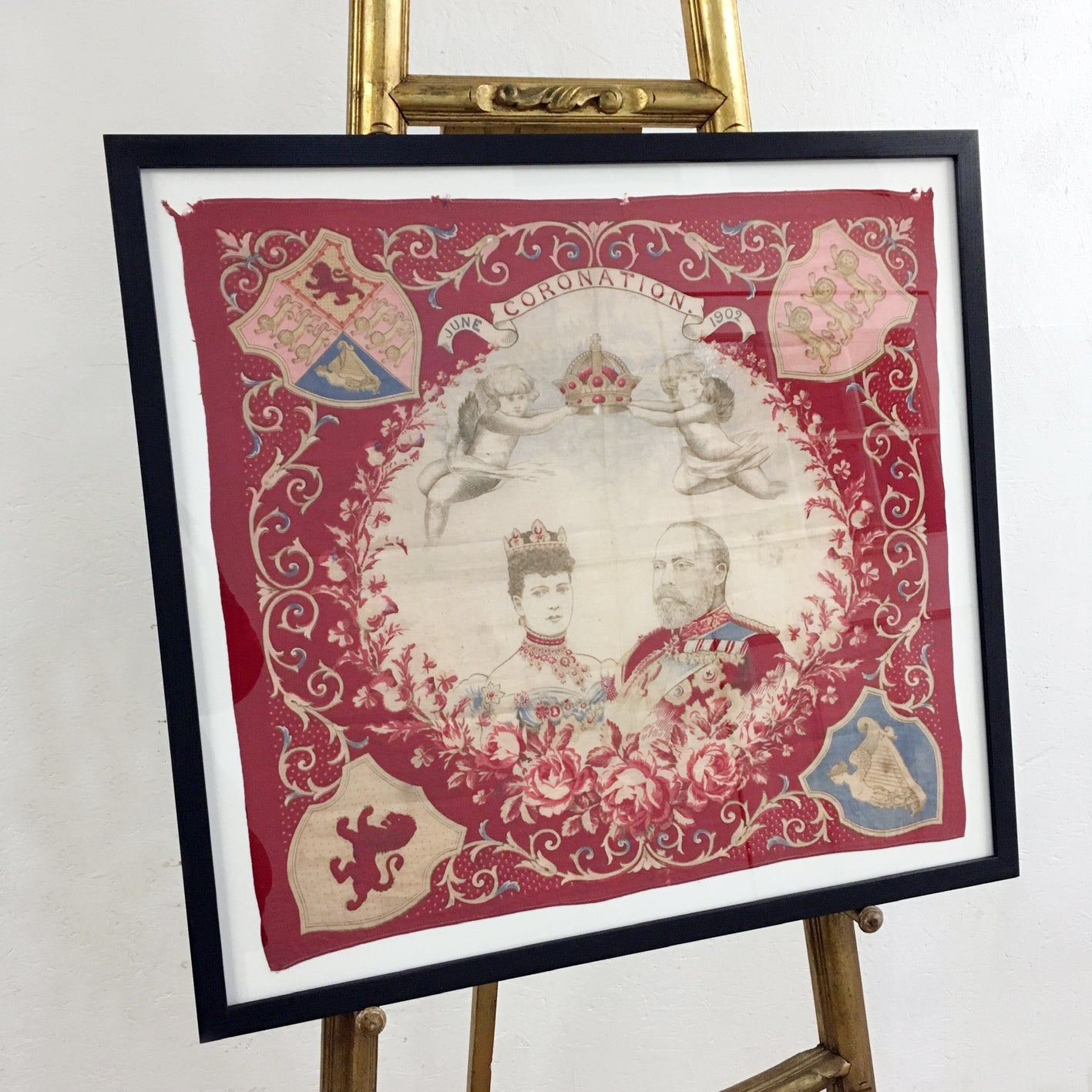 A framed antique embroidered textile artwork displayed on a gold-colored wooden easel. The square piece of embroidery features a red background with ornate gold floral scrollwork, cherubs, heraldic shields, and portraits of King Edward VII in the center.