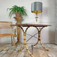 A vintage simulated bamboo table with a dark wood top, styled in a rustic room next to an arras armchair, a potted fern on a pedestal, and a table lamp with a gray and gold shade.